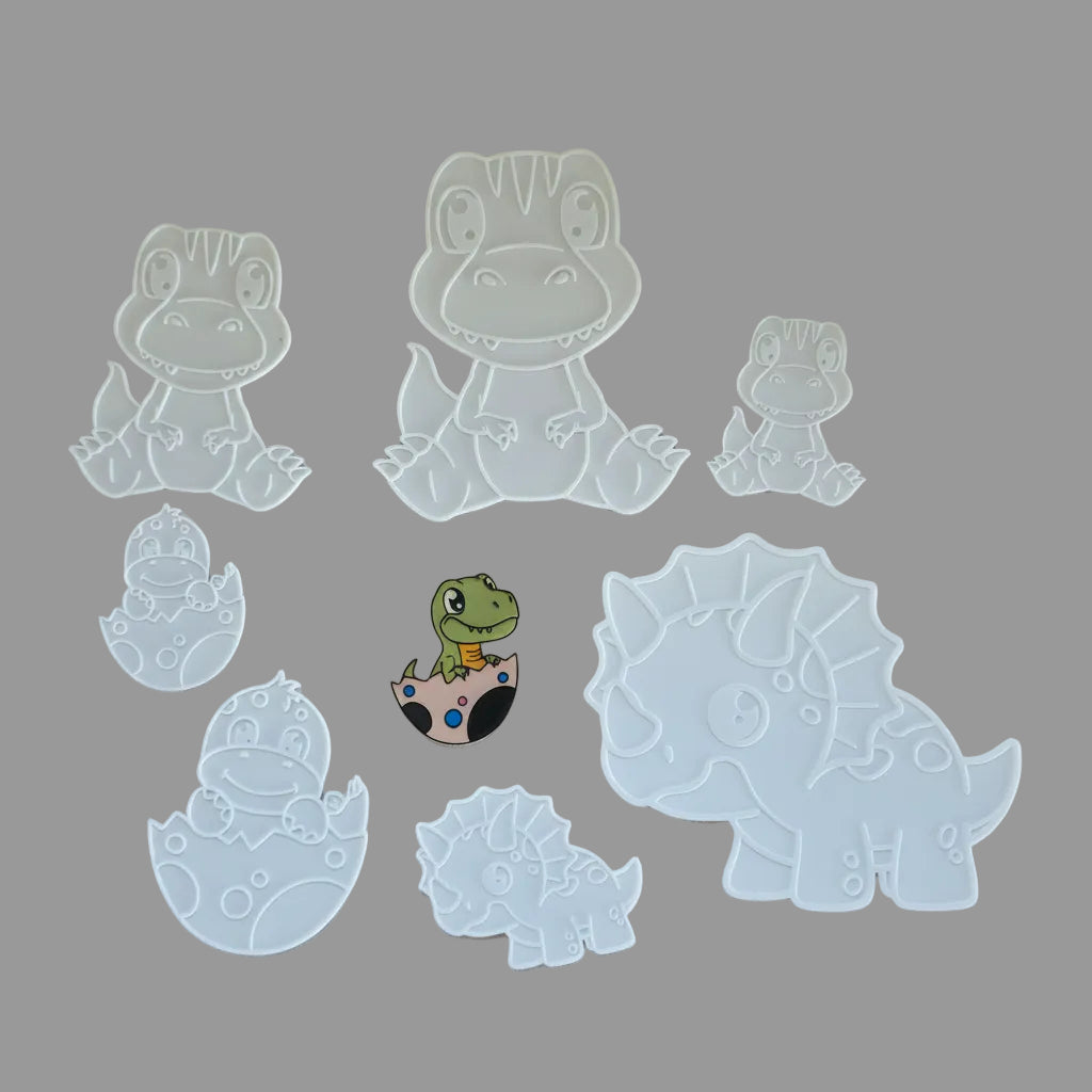 A set of white plastic dinosaur-themed stencils arranged on a white fabric background, including various dinosaur silhouettes and one small dinosaur hatching from an egg.